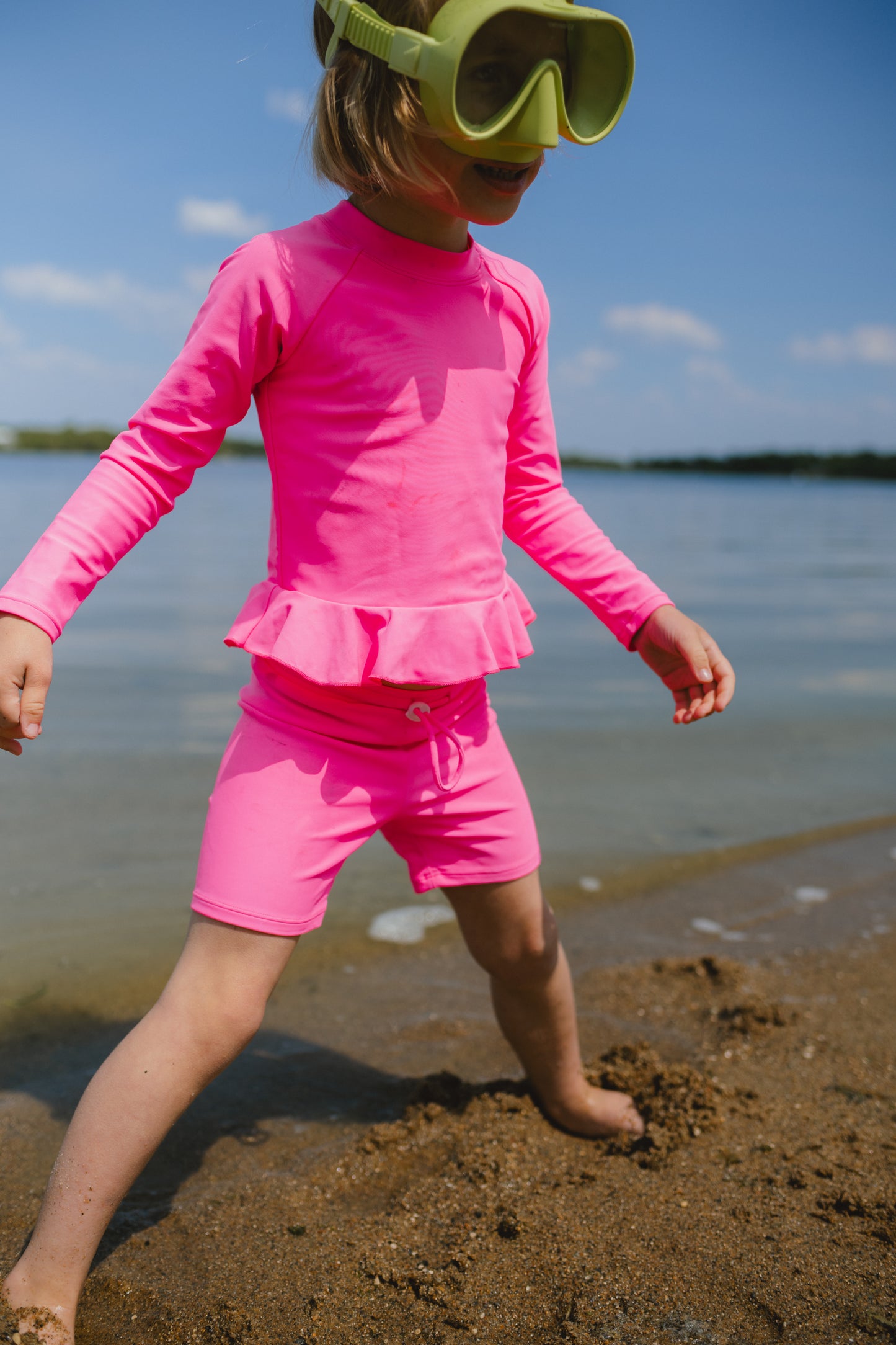 Child in pink rash guard and shorts standing on a beach with water and sky in the background