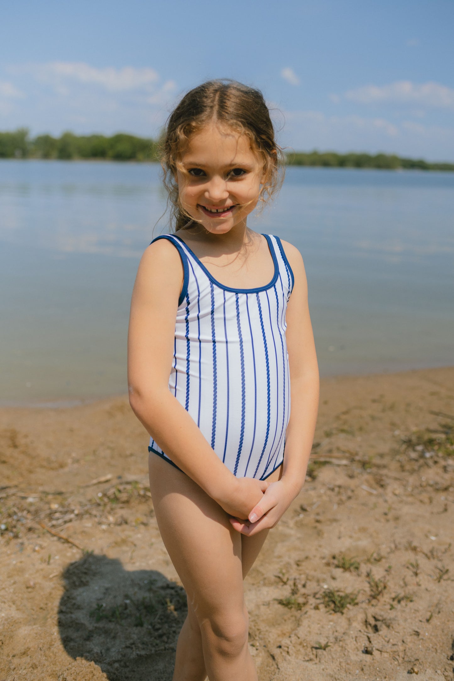 Young girl in a striped swimsuit standing on a sandy beach with water and sky in the background
