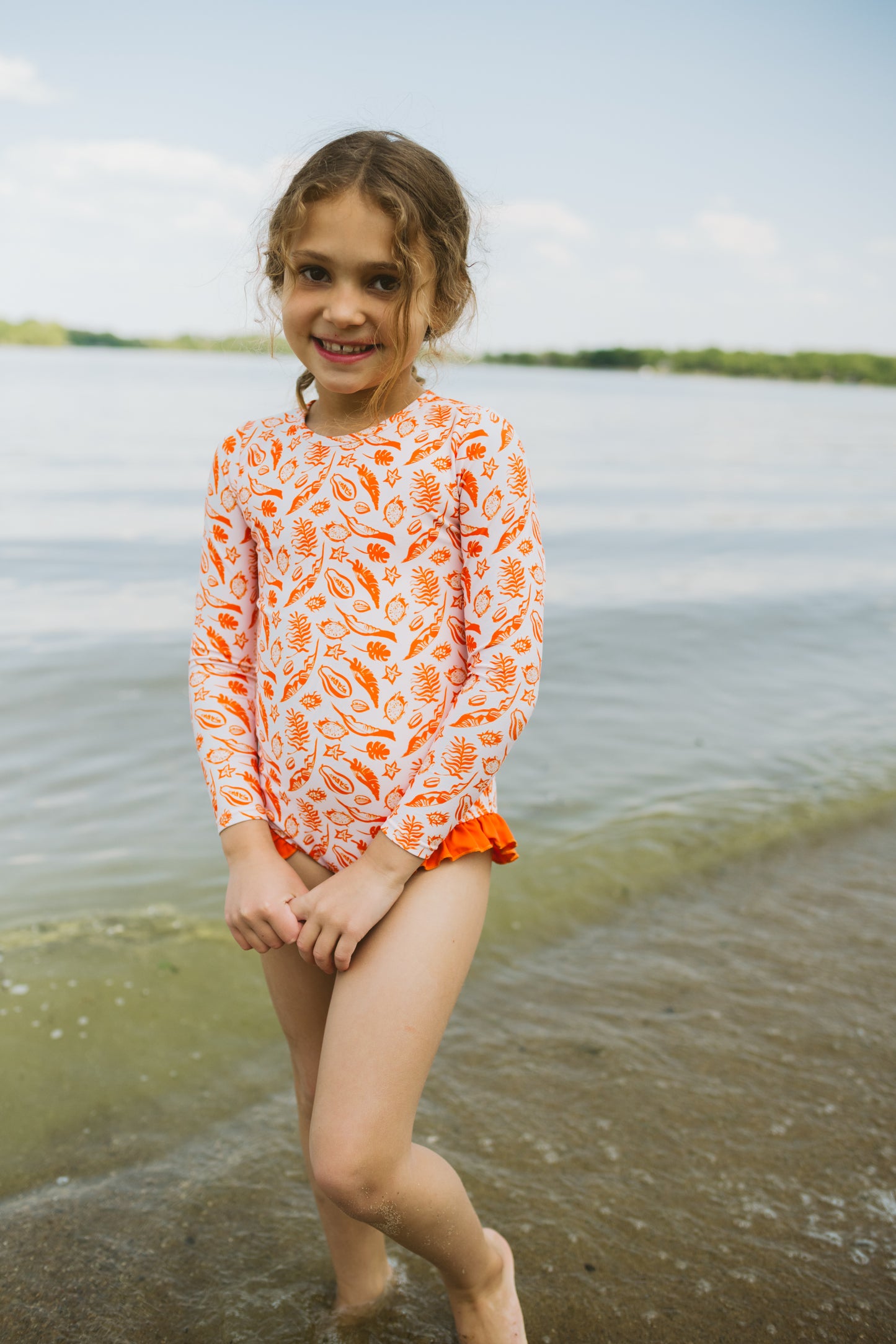 Young girl in an orange and white patterned shirt standing by a body of water.