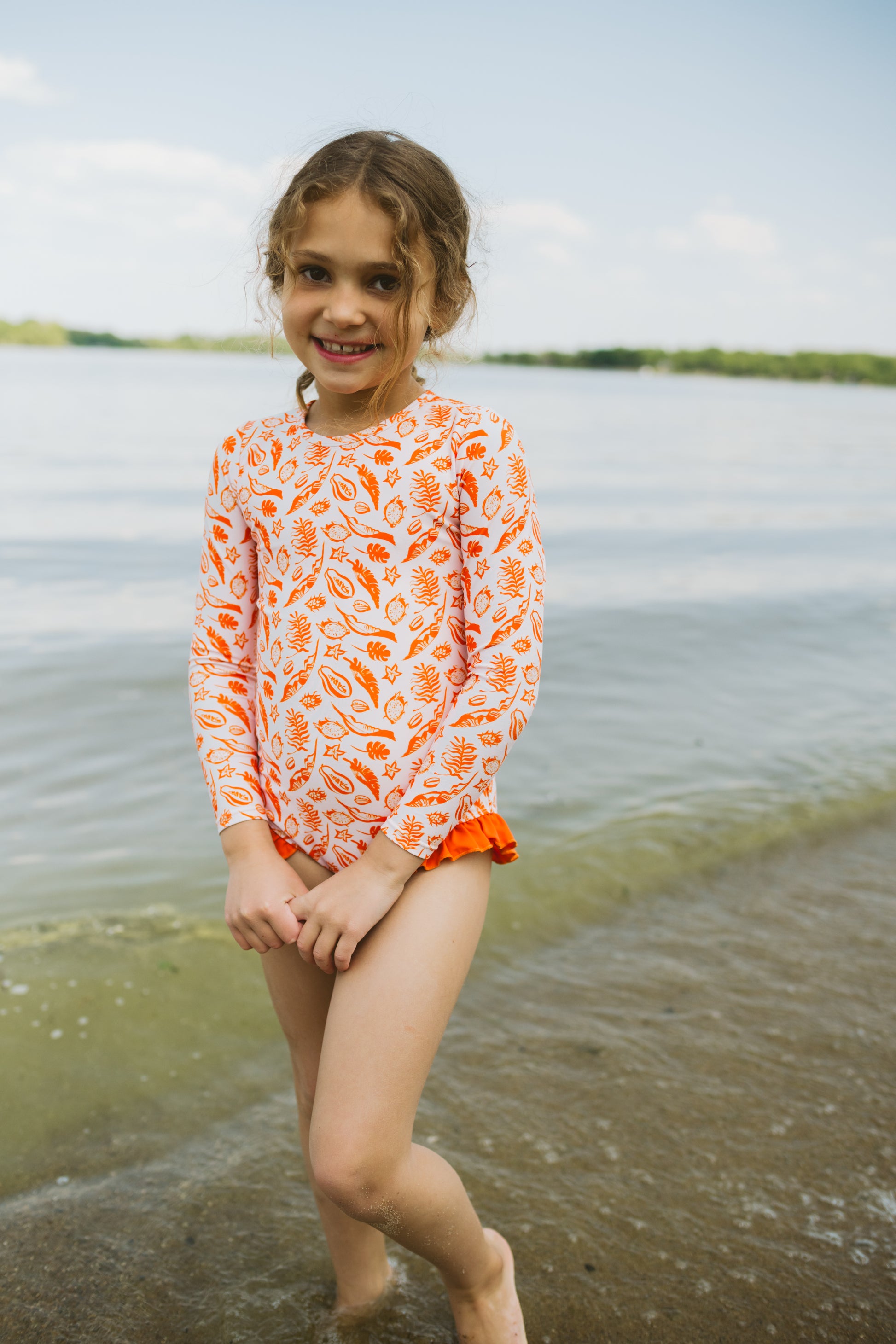 Young girl in an orange and white patterned shirt standing by a body of water.