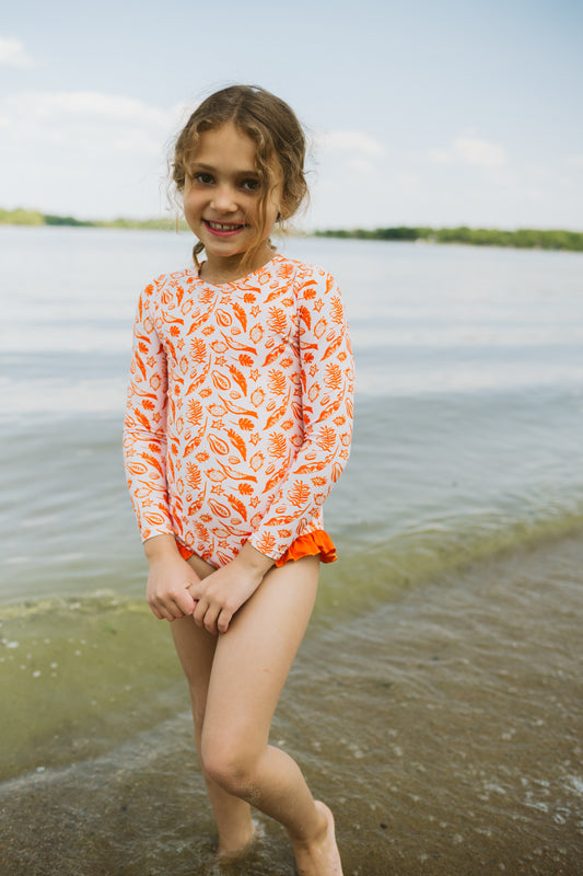 Young girl in an orange and white patterned shirt standing by a body of water.