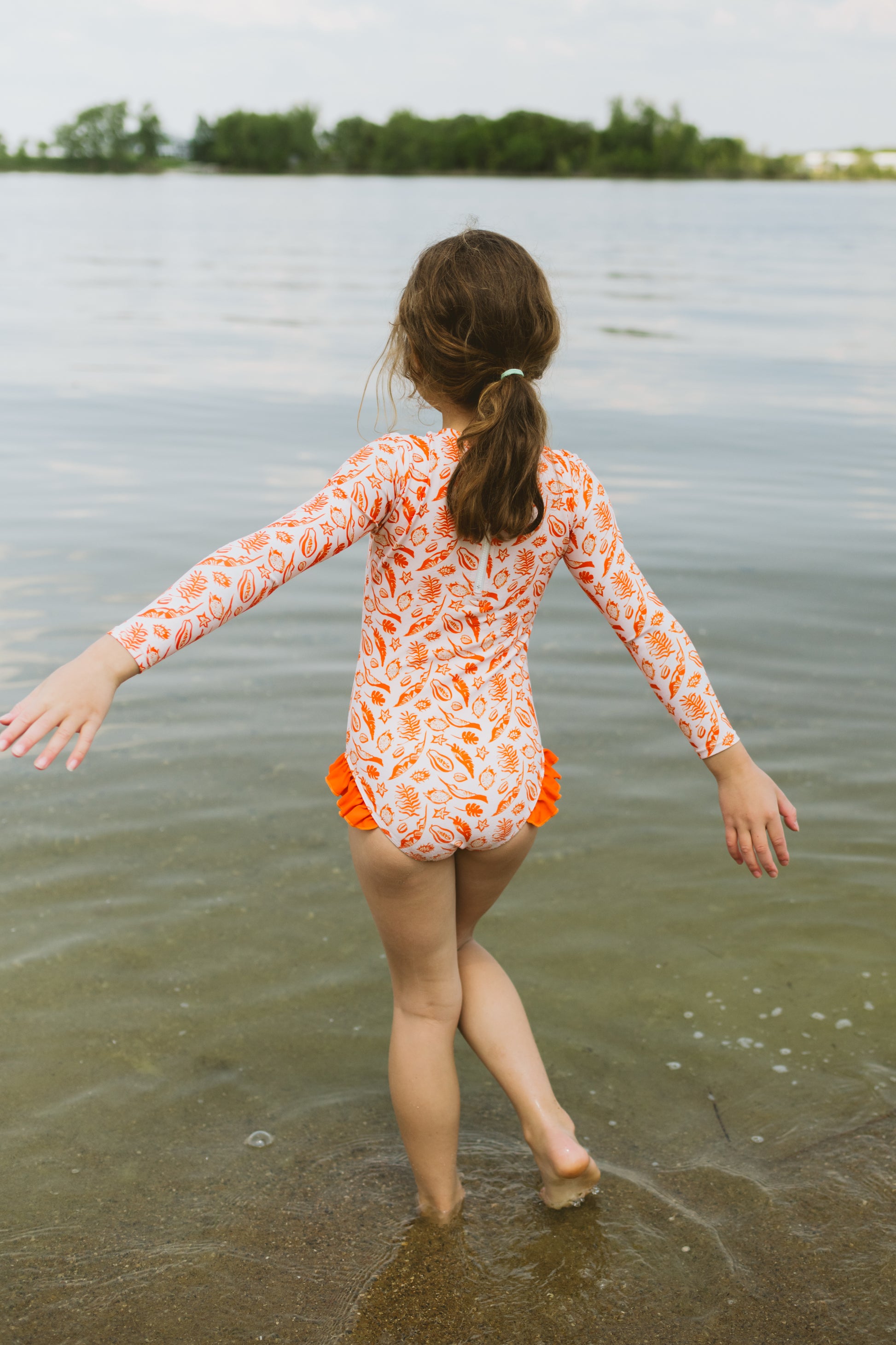 Child in an orange and white long sleeve swimsuit standing in shallow water by a lake.
