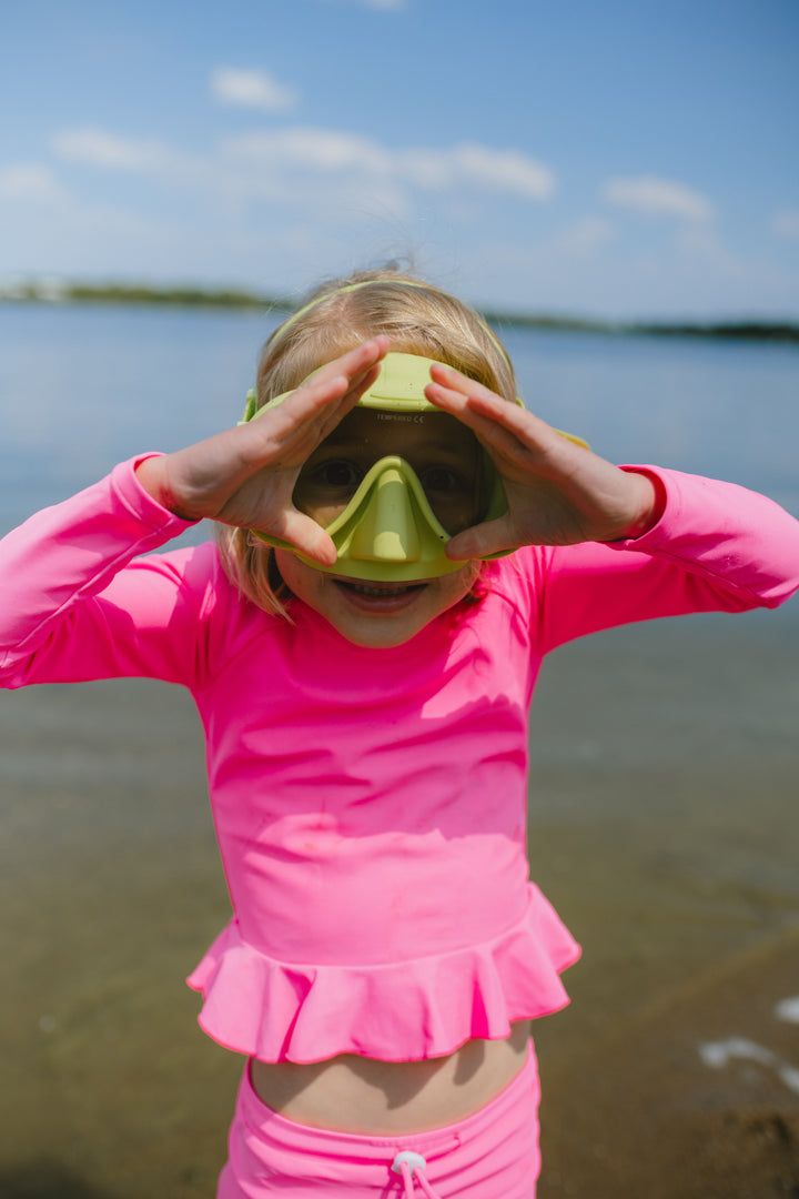 Child wearing a pink swimsuit and green goggles by a body of water.