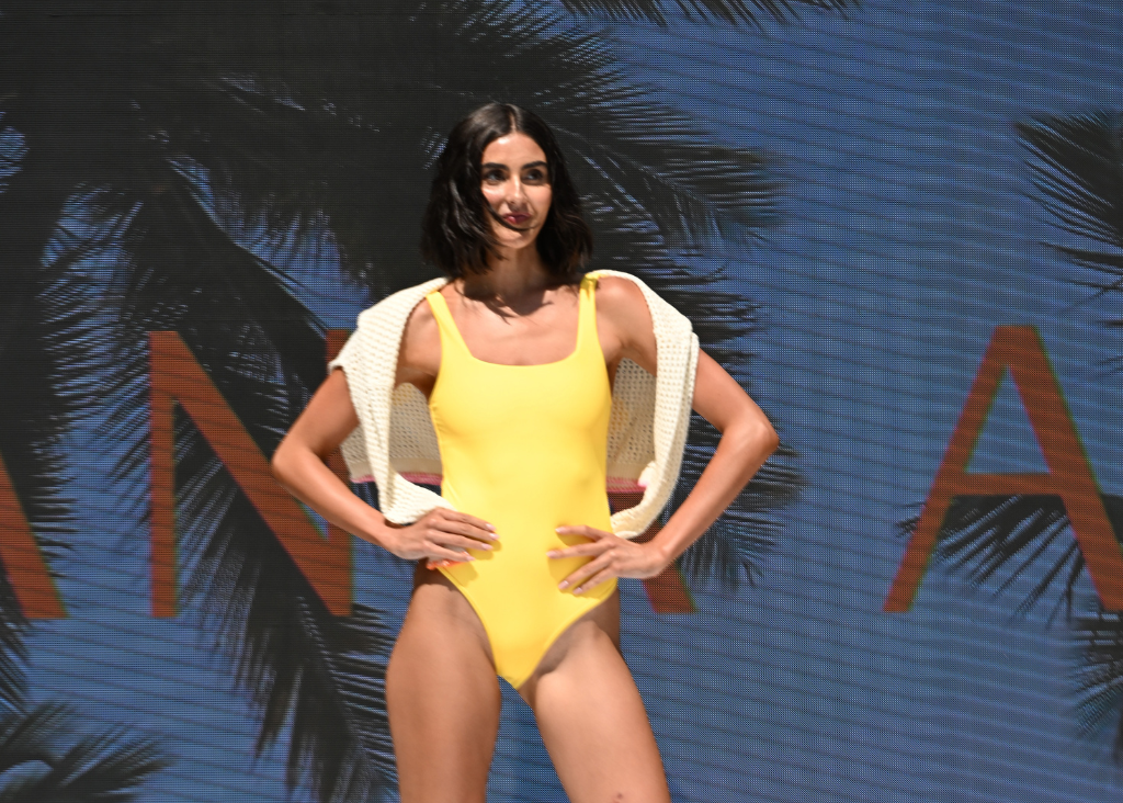 A model walks a fashion runway wearing a bright yellow one-piece swimsuit with a white draped jacket over her shoulders, set against a blue background with palm tree silhouettes and partial orange lettering.