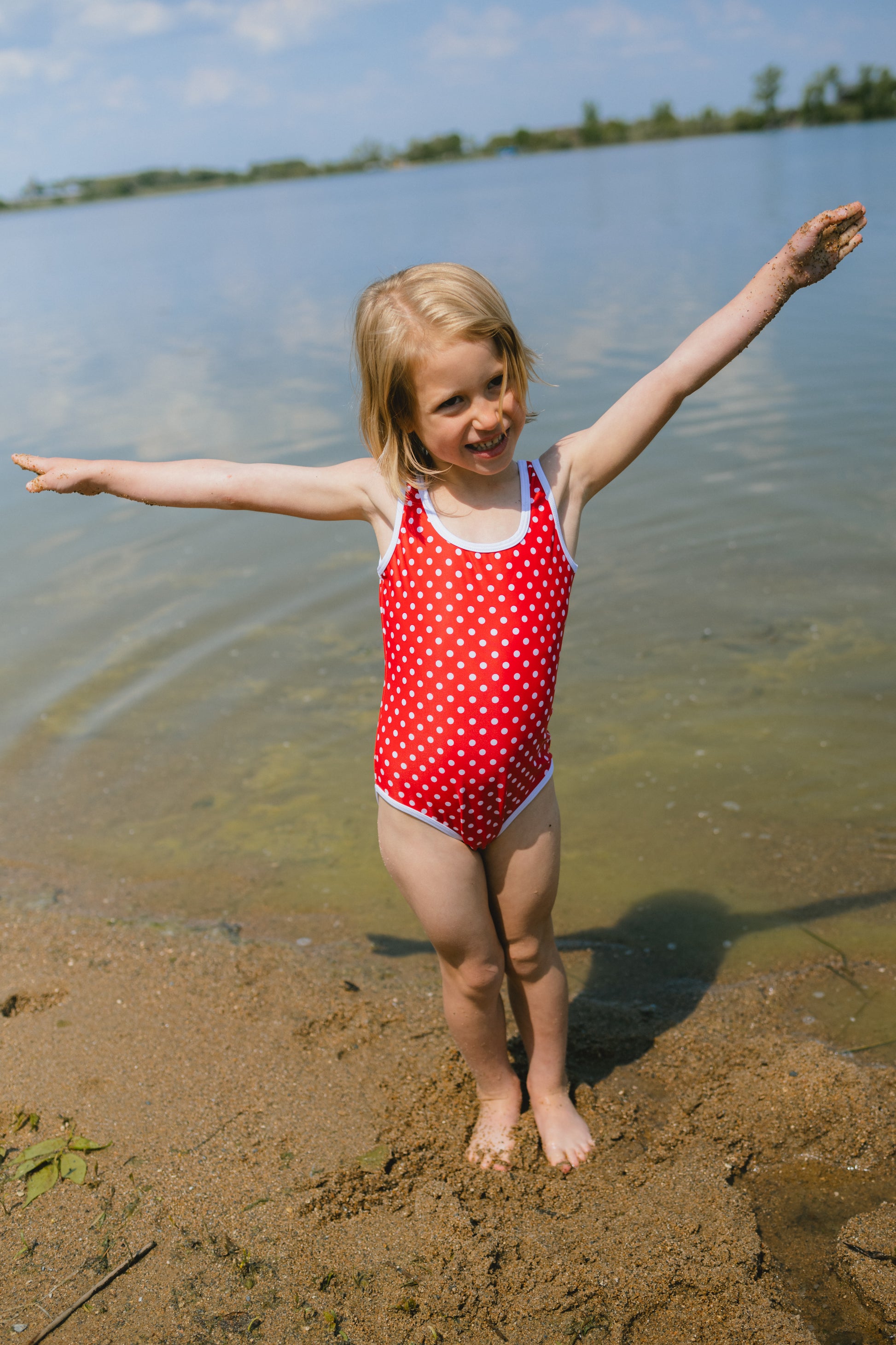 Young girl in a red polka dot swimsuit standing on a sandy beach with arms outstretched.