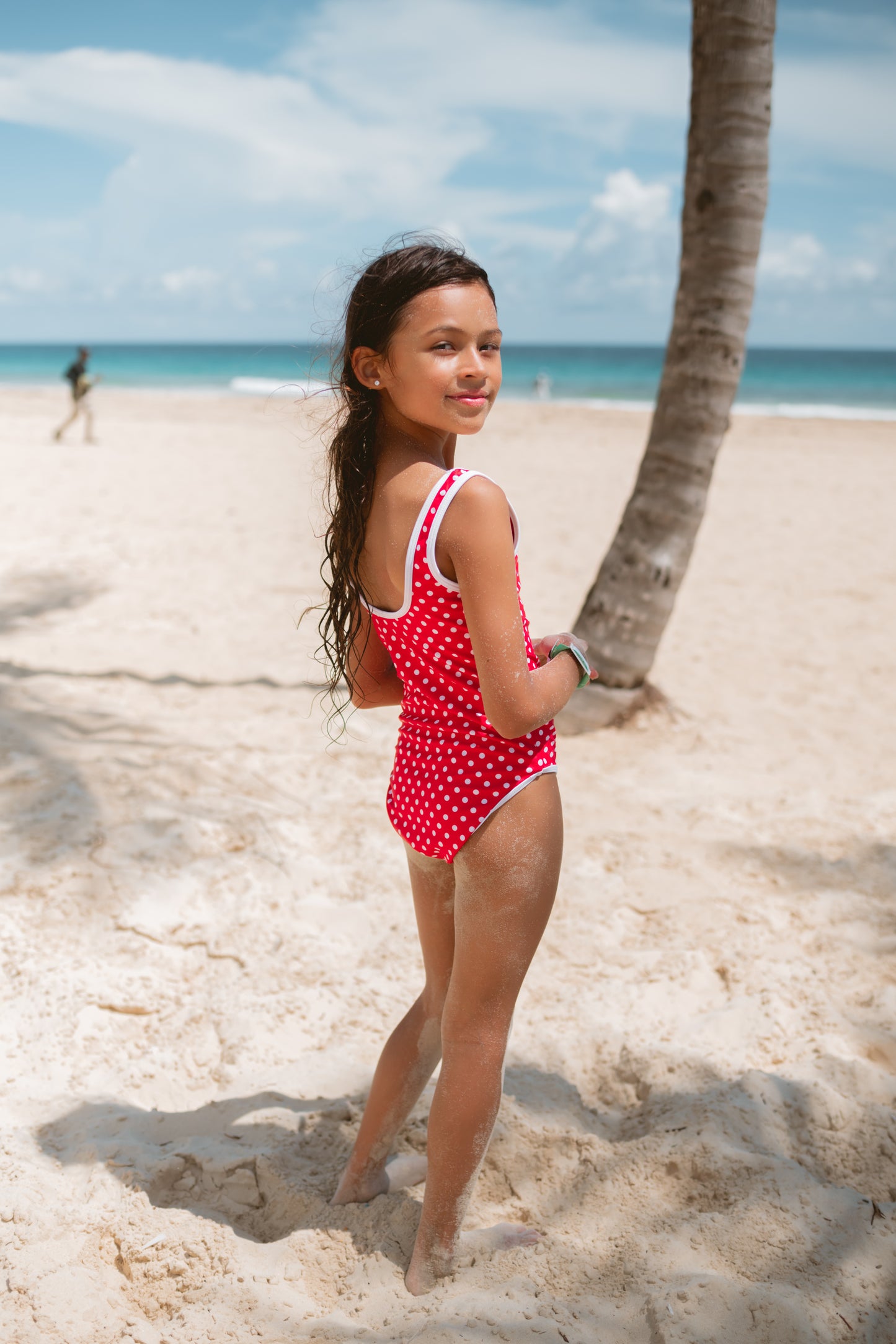 Young girl in a red polka dot swimsuit standing on a sandy tropical beach with a palm tree and ocean in the background.