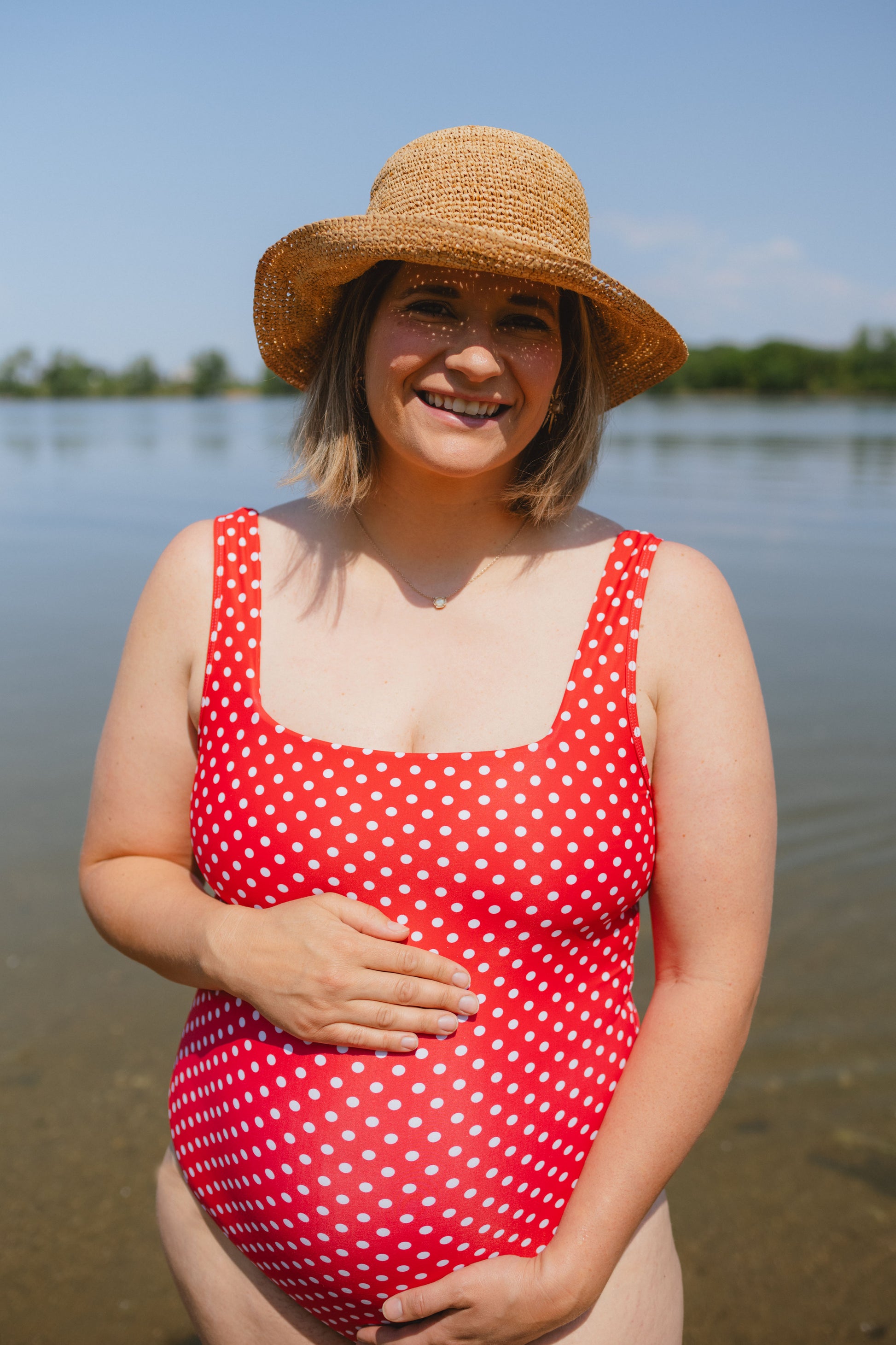 Pregnant woman in a red polka dot swimsuit and straw hat standing by a body of water.