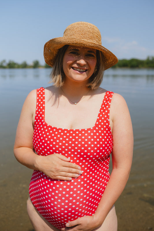 Pregnant woman in a red polka dot swimsuit and straw hat standing by a body of water.